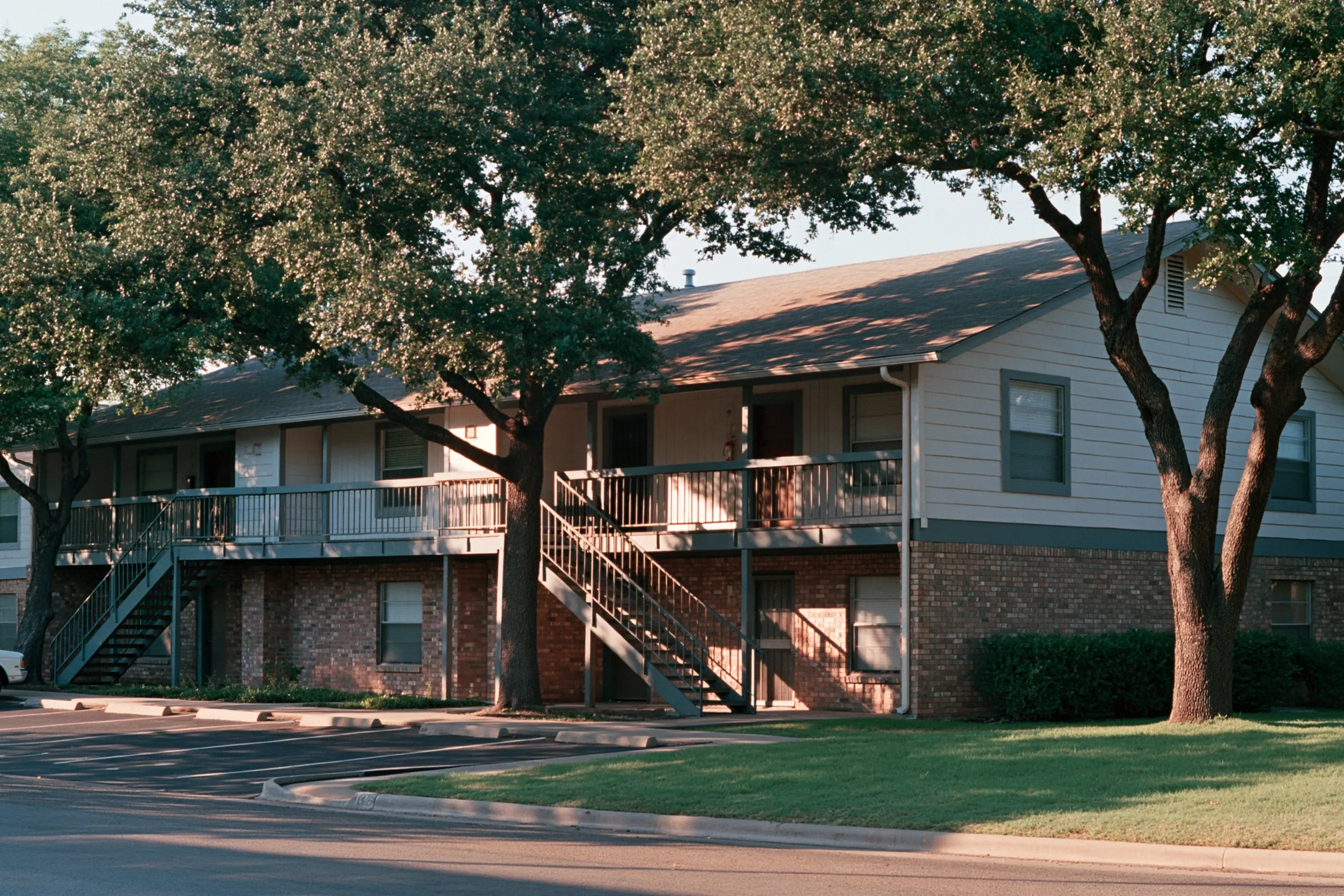 Two-story 1980s garden apartment building with exterior walkways, painted siding above brick, under live oaks